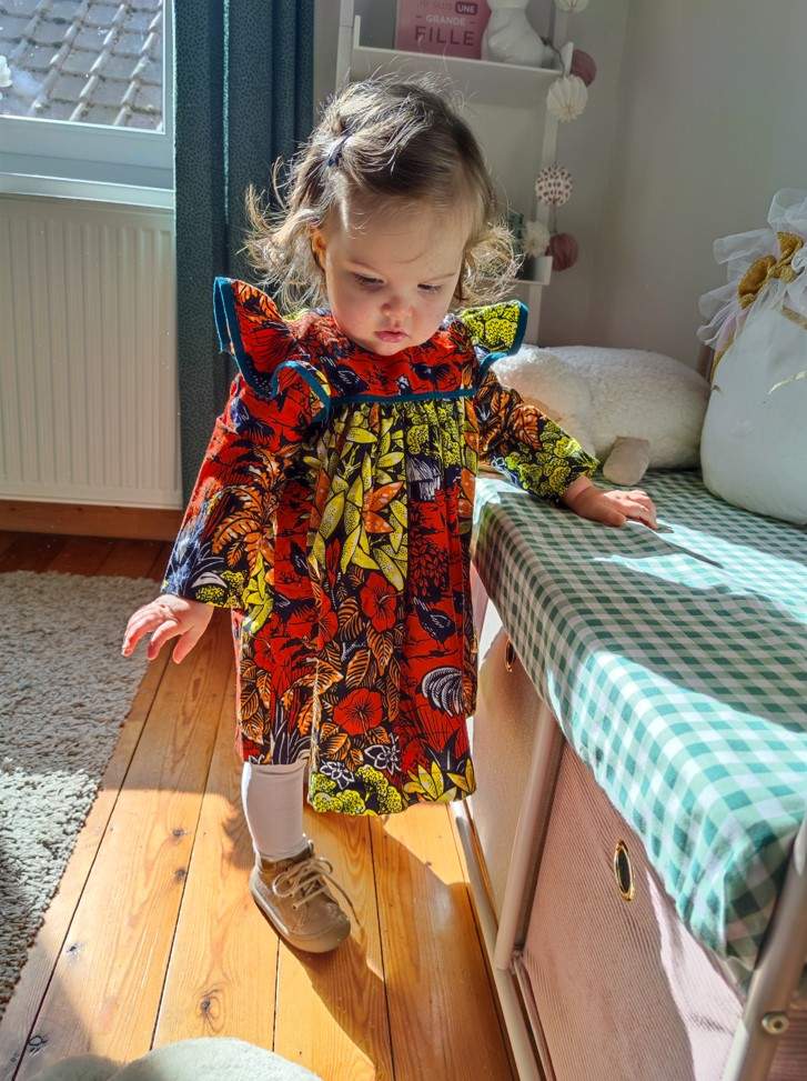 Bébé fille debout qui regarde par terre, elle s'appuie sur un banc recouvert de tissu imprimé vichy