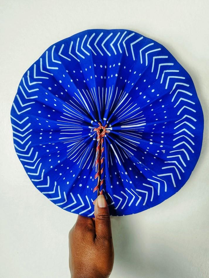 éventail en wax tenu à la main, les motifs de ce wax sont en forme de chevrons blancs sur fond bleu, il forme un cercle concentrique par des petits points et traits. Le fond de la matière est bleu indigo, c'est un motif typiquement africain