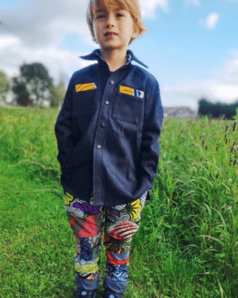 garçon blond vu de face portant une chemise ample en jeans sur un pantalon multicolore, il se tient debout au milieu de l'herbe verte