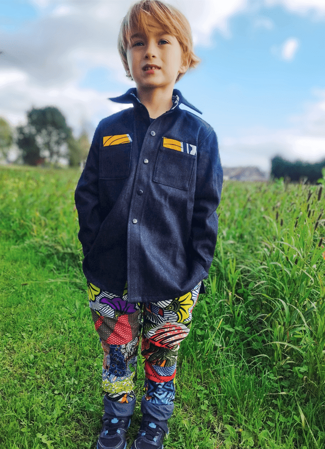 garçon blond vu de face portant une chemise ample en jeans sur un pantalon multicolore, il se tient debout au milieu de l'herbe verte