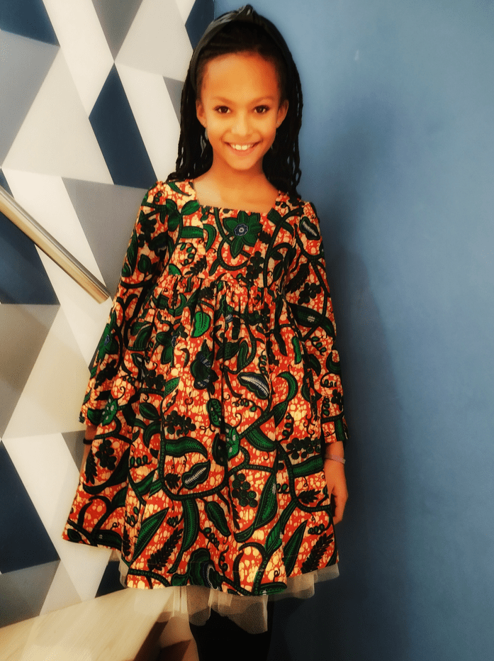 fille debout portant une robe en tissu wax imprimé de motifs de feuilles vertes sur un fond orange brique