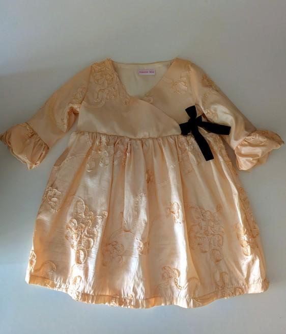 robe en taffetas brodée pour fille, posée à plat, il y a un nœud noir sur le côté, la robe est couleur champagne