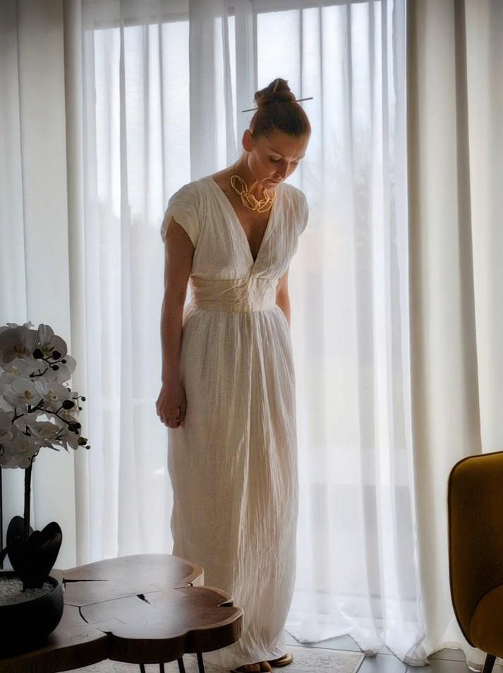 Femme à la tête baissée, elle porte une longue robe blanche. La robe est resserrée à la taille et les épaules sont larges et retombent sur les bras