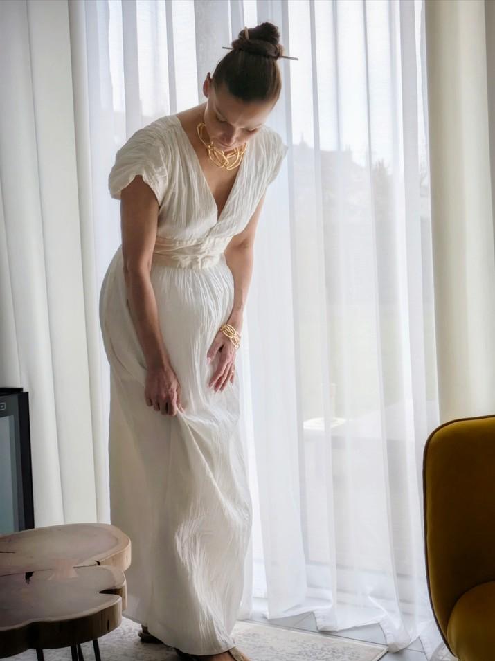 femme debout devant une fenêtre, elle porte une robe blanche et redresse le pan de sa robe